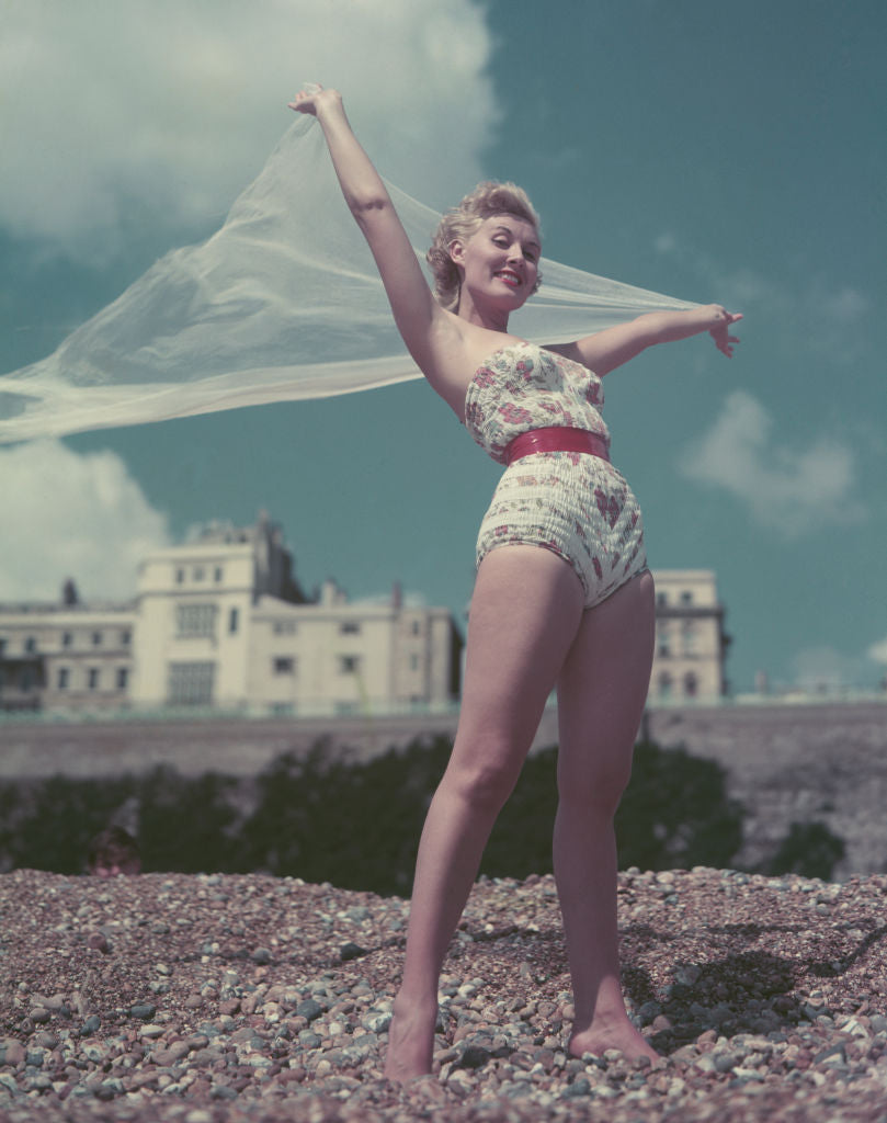 Colour photograph of Jill Day in a floral swimsuit on a pebble beach, holding a sheer scarf above her head in the breeze, circa 1955