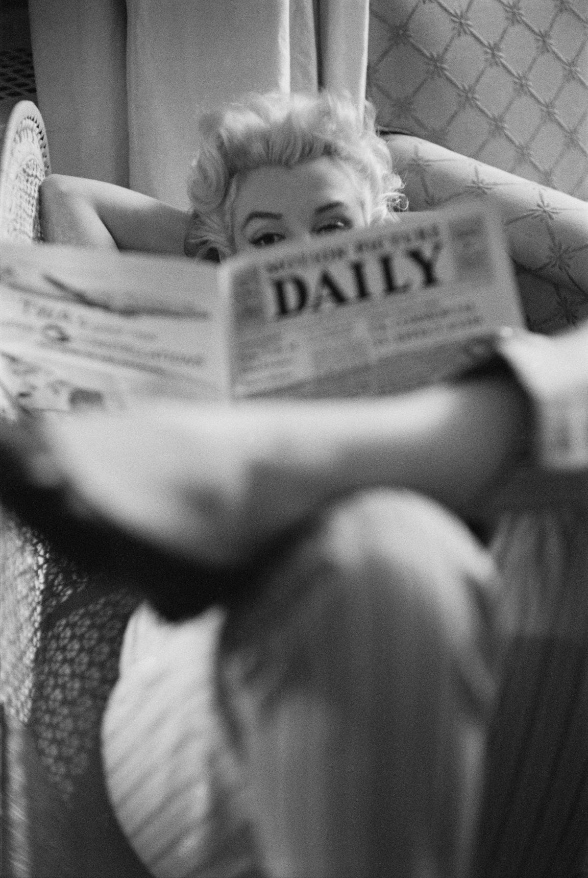 Black and white photograph of Marilyn Monroe peeking over a copy of Motion Picture Daily with her legs crossed in the foreground, New York, 1955
