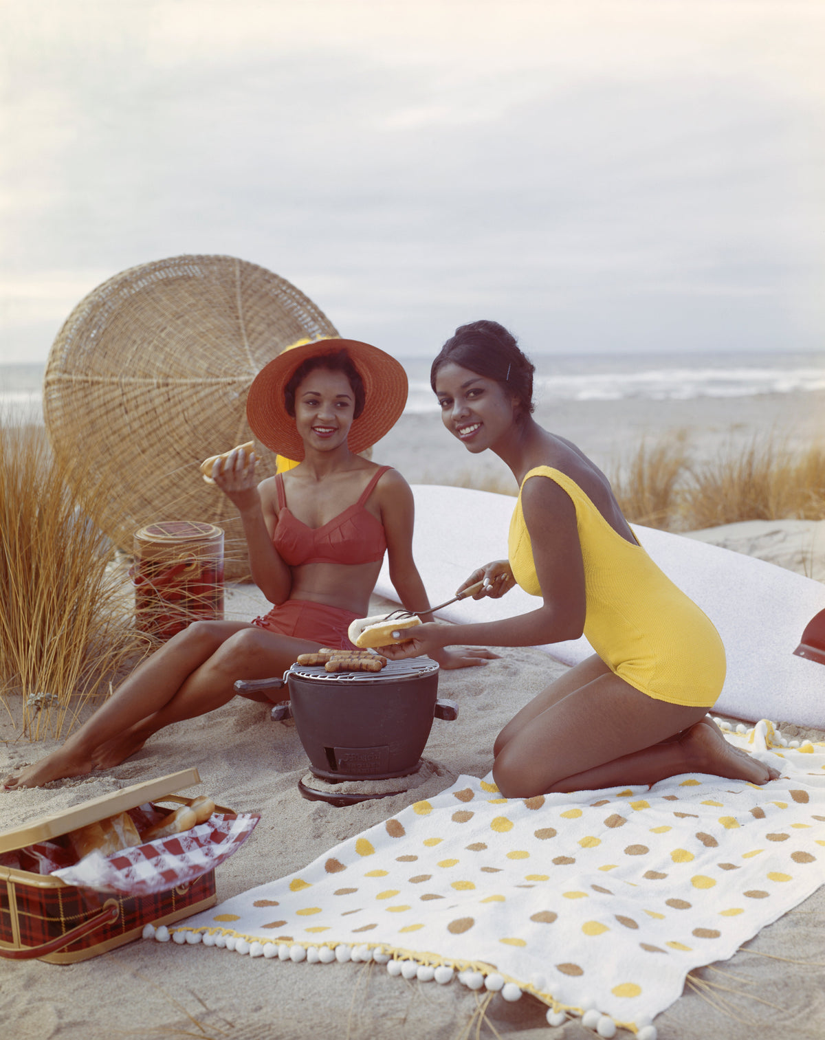 Colour photograph of two smiling young women in swimsuits having a picnic on a polka dot blanket on the beach