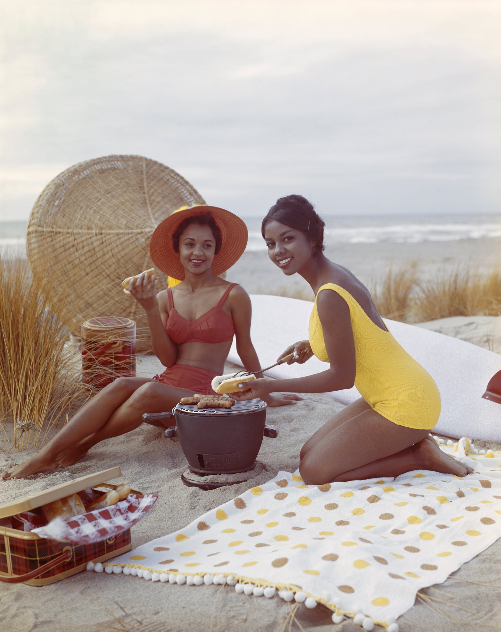 Colour photograph of two smiling young women in swimsuits having a picnic on a polka dot blanket on the beach