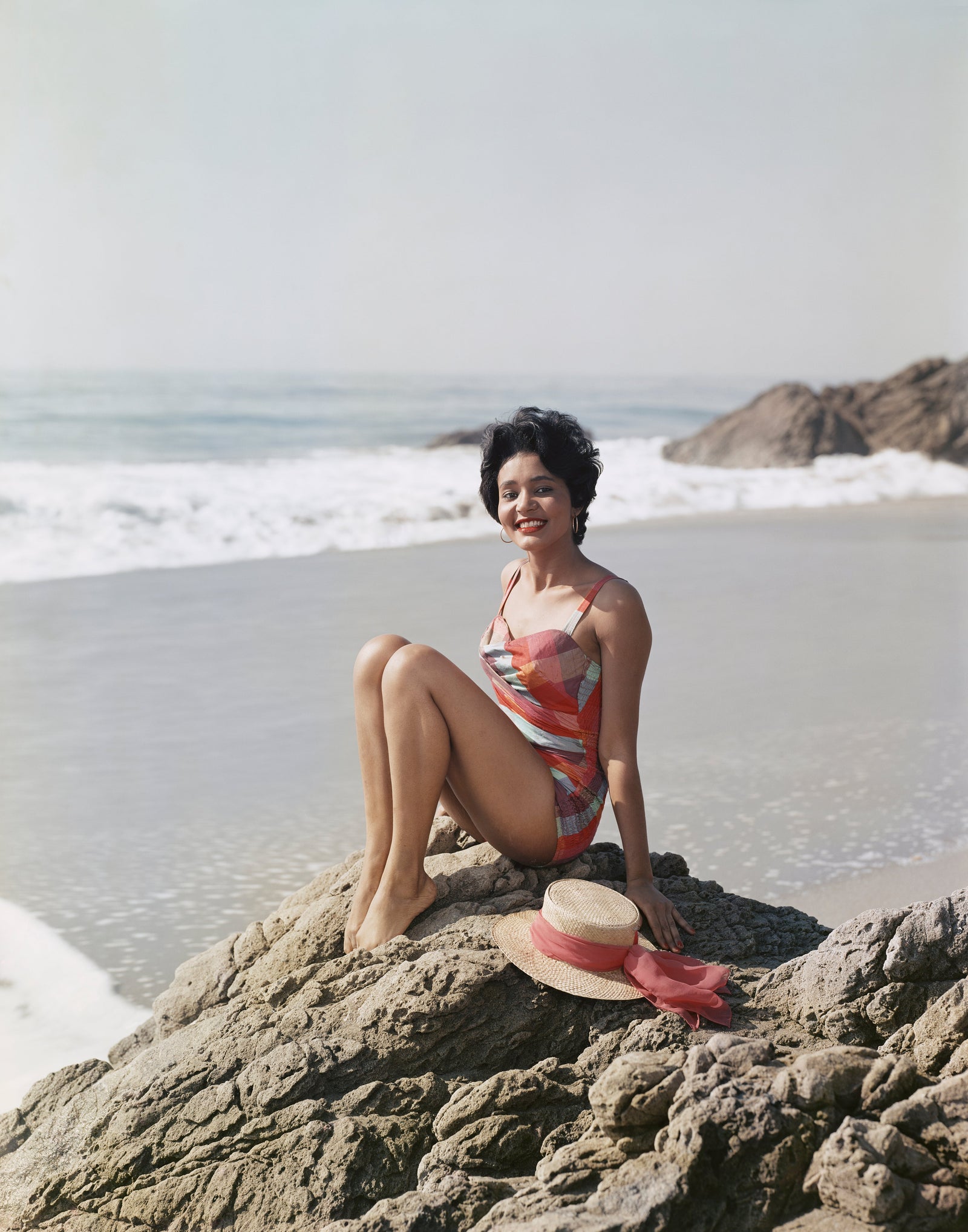 Colour photograph of a smiling young woman in a swimsuit sitting on a rock by the sea with a straw hat beside her