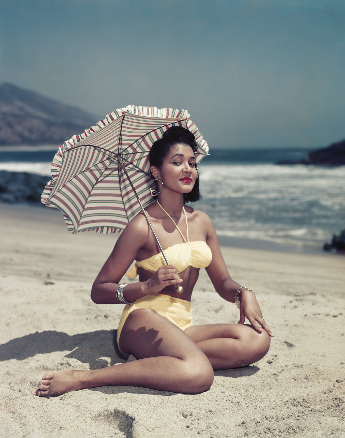 Colour photograph of a smiling woman in a yellow bikini sitting on the sand beneath a striped parasol by the sea