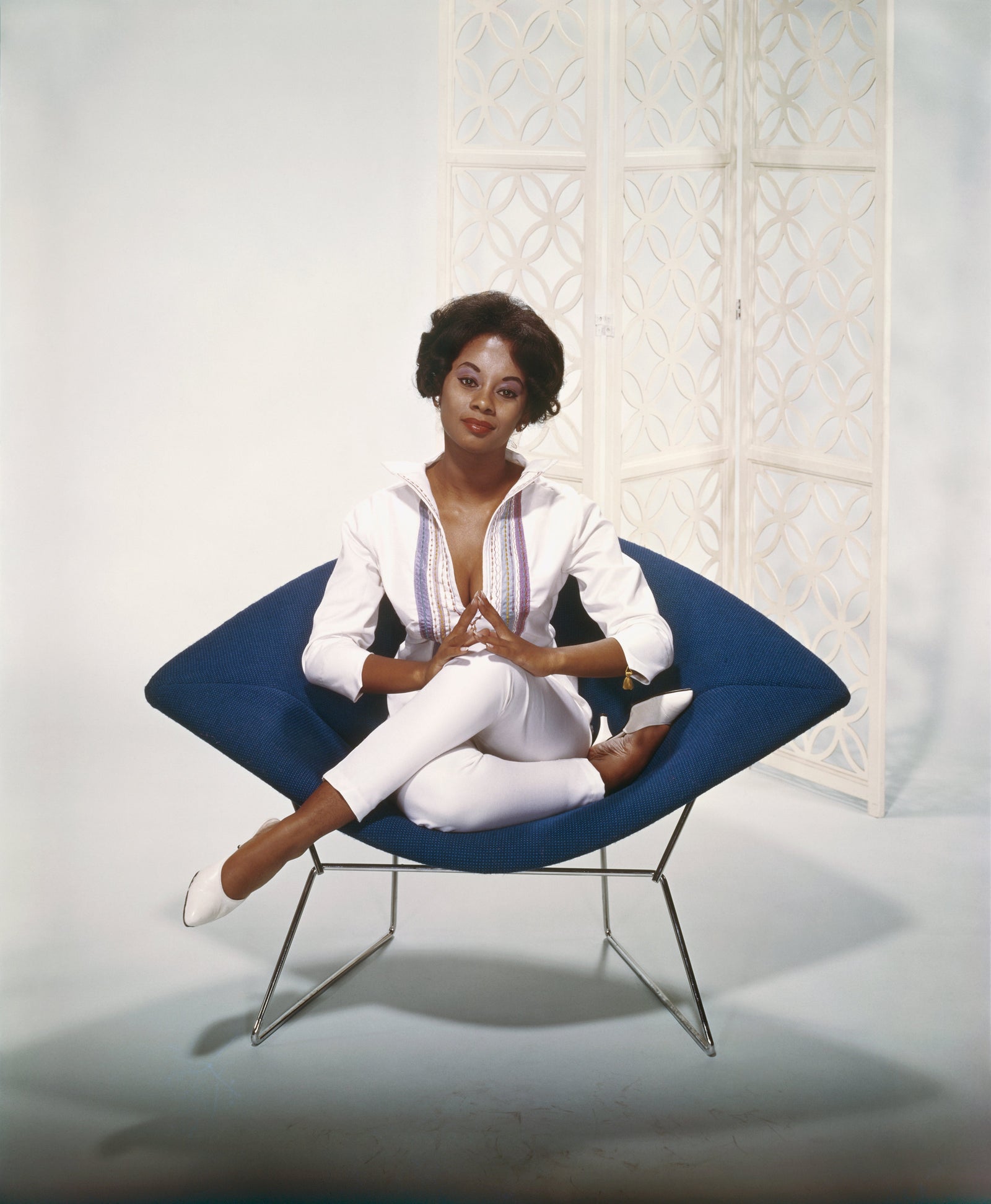 Colour photograph of a smiling young woman dressed in white sitting cross-legged in a blue wire-framed lounge chair