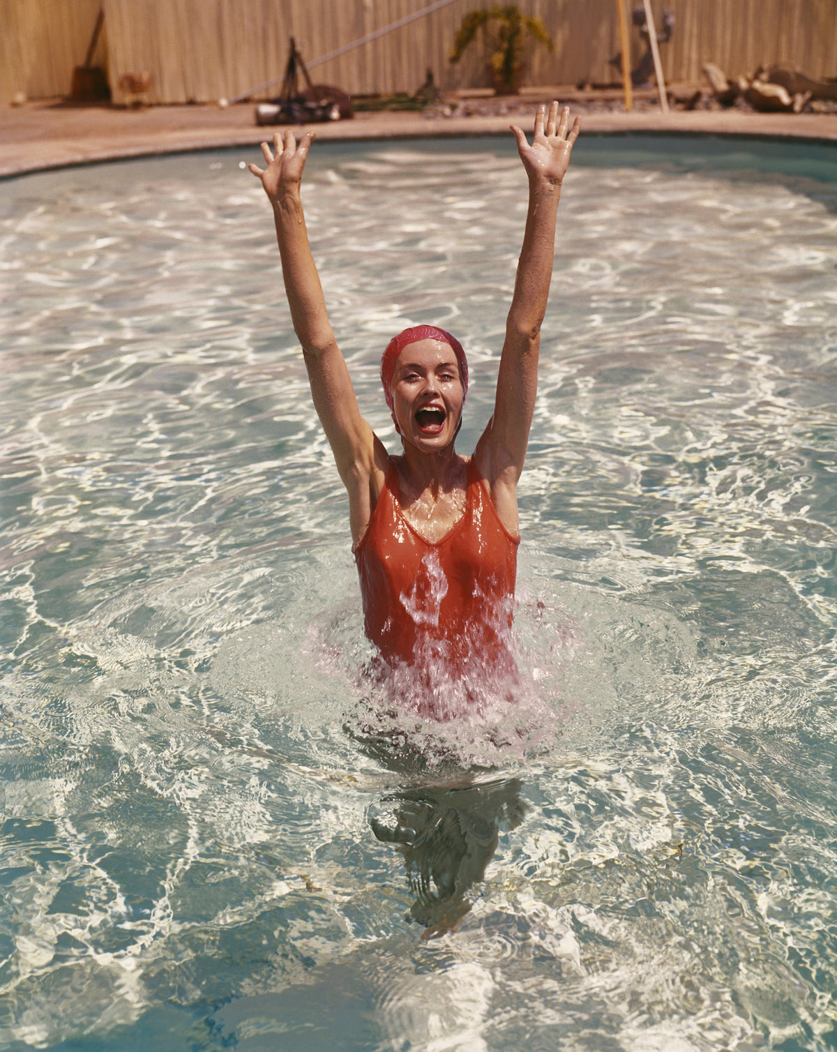 Colour photograph of a young woman in a red swimsuit and swim cap rising from a sunlit swimming pool with arms raised