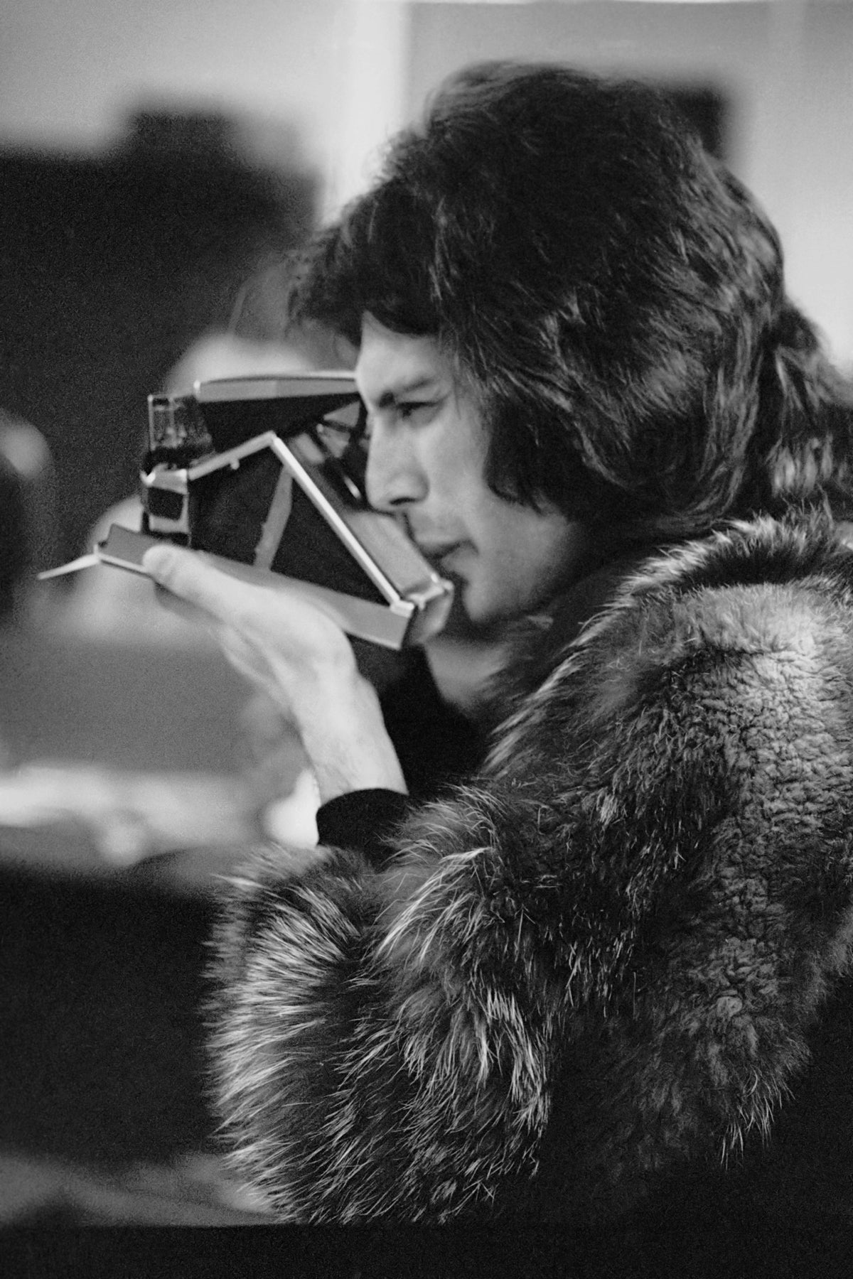 Black and white photograph of Freddie Mercury in a fur coat taking a picture with a Polaroid camera, Los Angeles, circa 1977