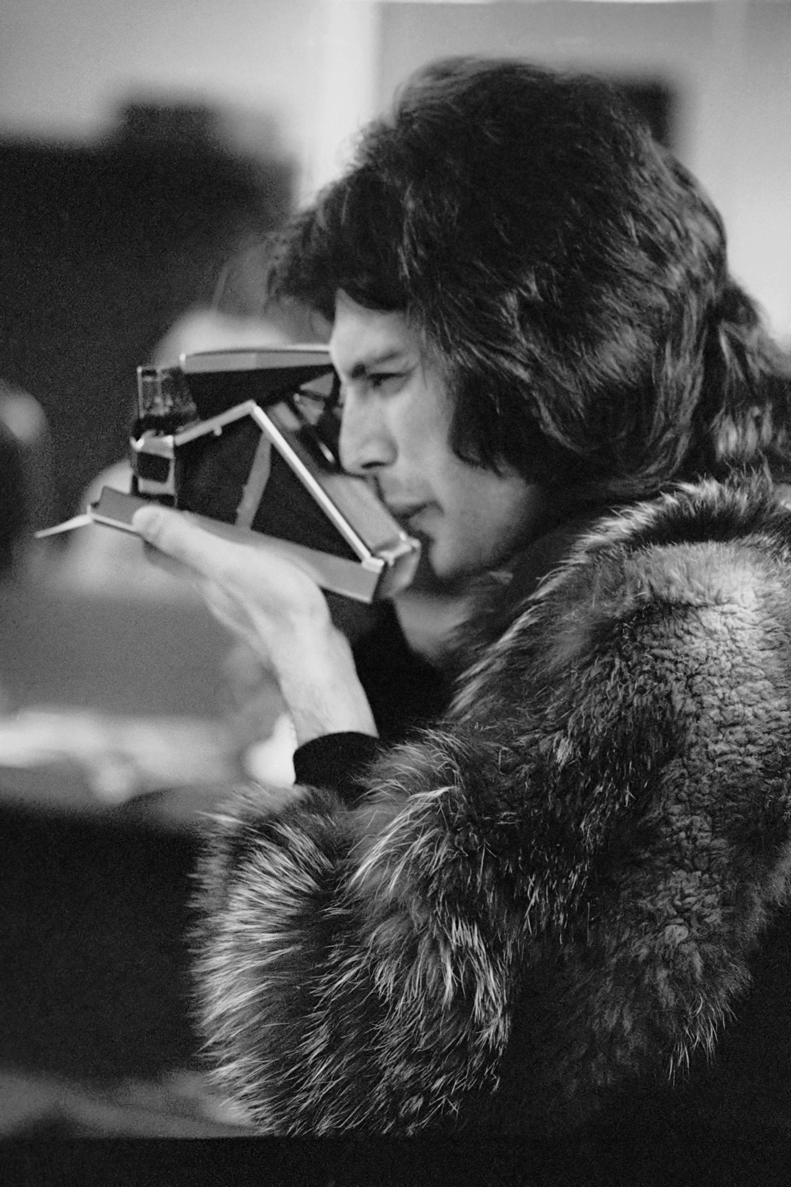 Black and white photograph of Freddie Mercury in a fur coat taking a picture with a Polaroid camera, Los Angeles, circa 1977