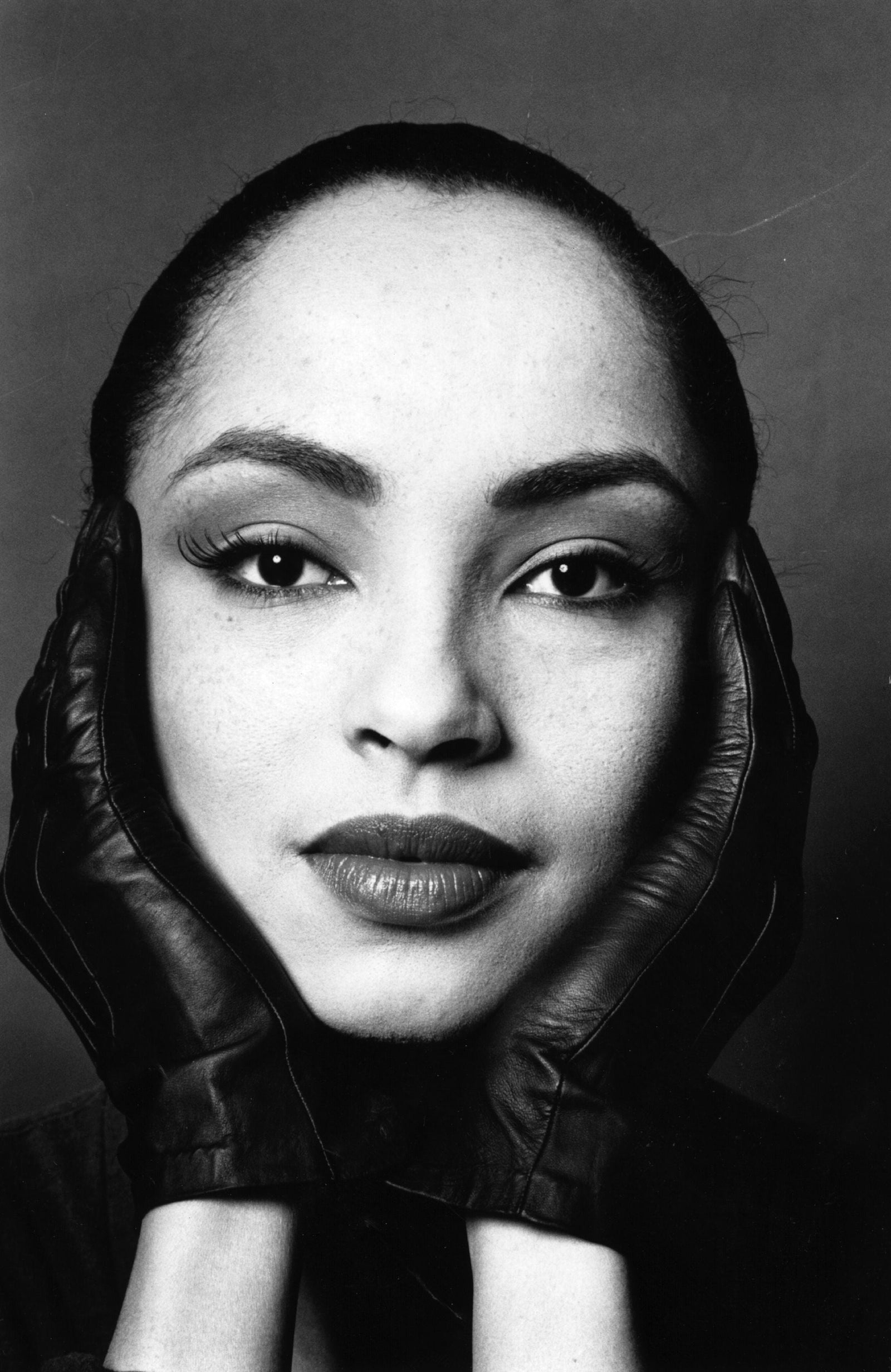 Black and white close-up portrait of Sade with her face framed by black leather gloved hands and a headscarf