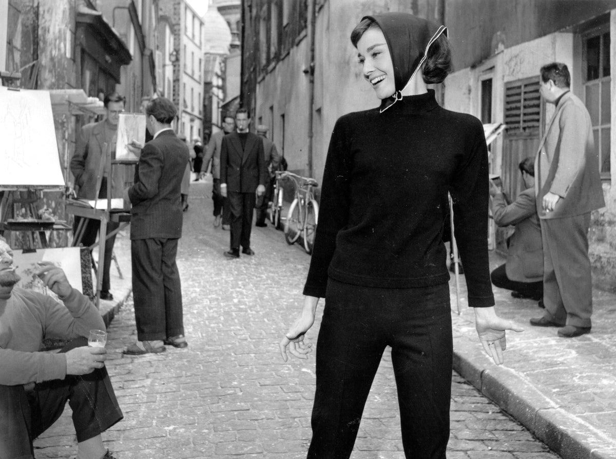Black and white photograph of Audrey Hepburn in a black jumper and headscarf on a cobbled Paris street while filming Funny Face, 1956