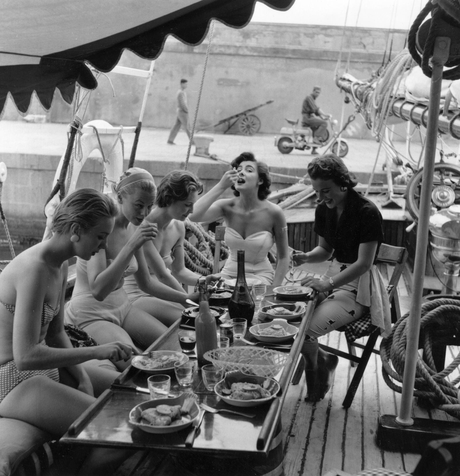 Black and white photograph of five models in swimwear eating lunch on the deck of a yacht on the Cote d'Azur, 1955