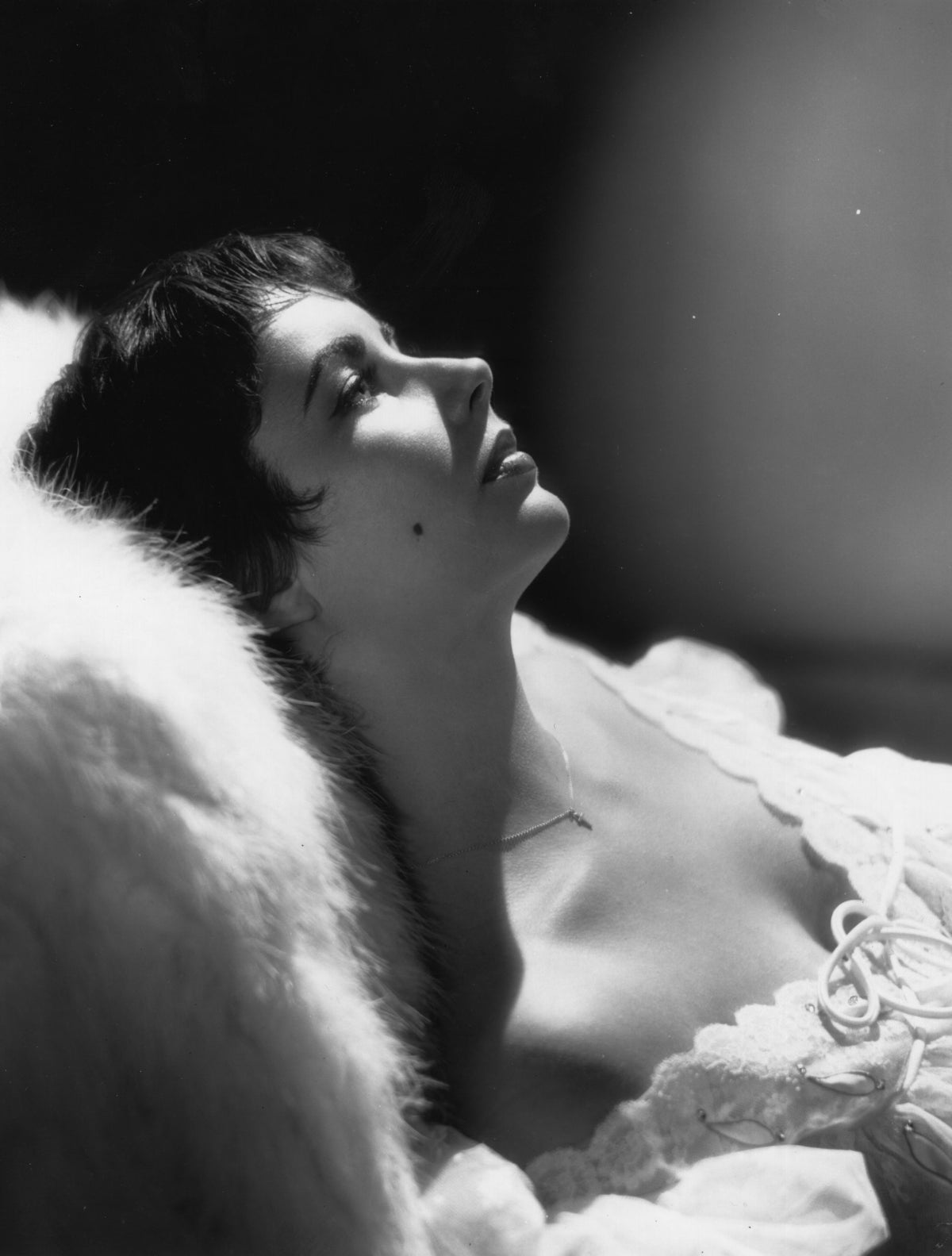 Black and white photograph of Elizabeth Taylor reclining in bed, her face in profile as she gazes upwards