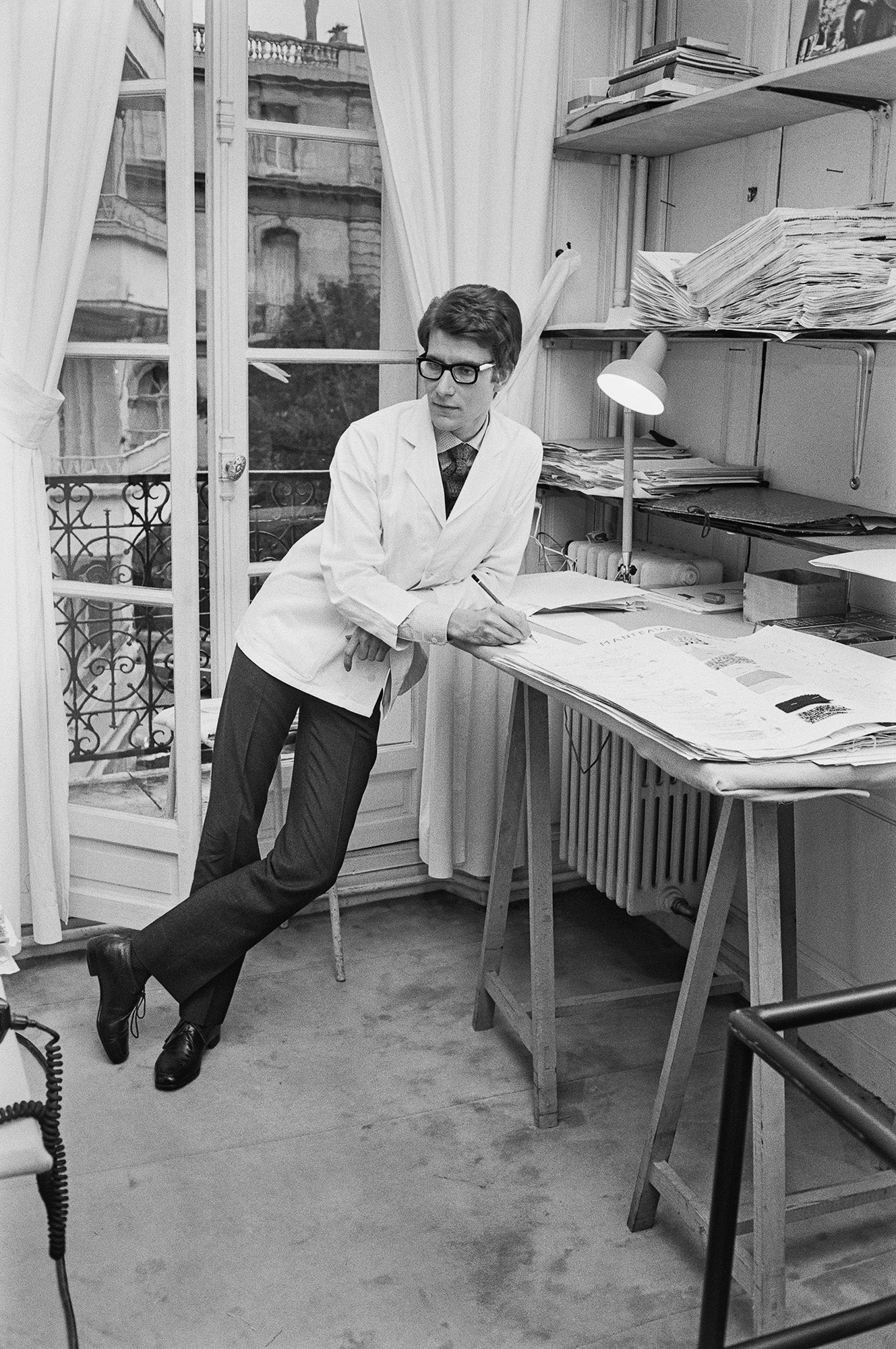 Black and white photograph of Yves Saint Laurent in a white coat leaning on a drawing table in his Paris studio, 1965