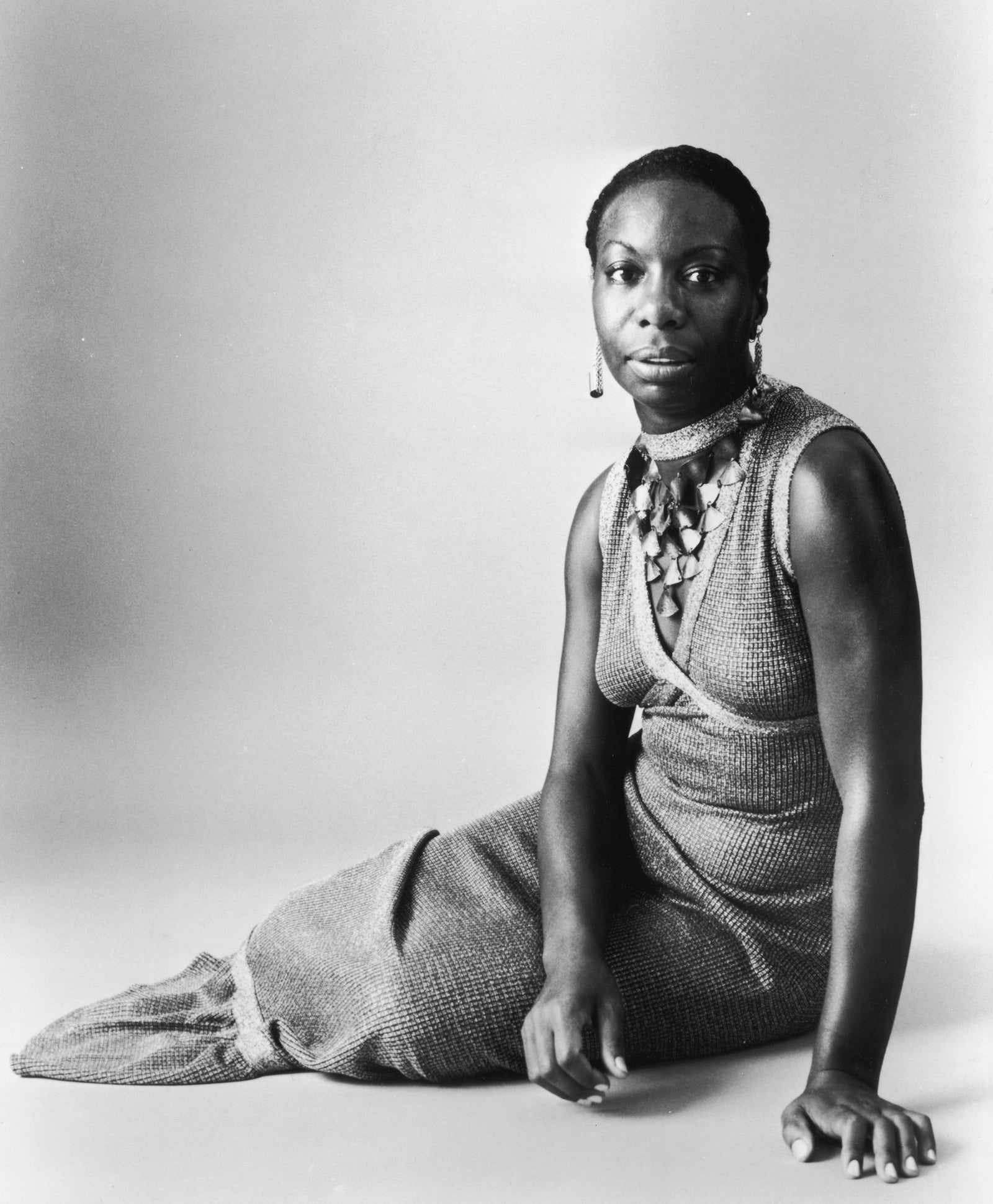 Black and white studio portrait of Nina Simone seated on the floor in a sleeveless dress and shell necklace, circa 1968