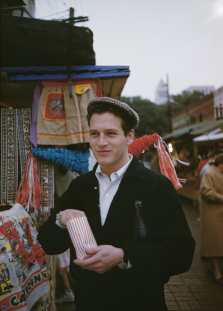 Colour photograph of Paul Newman in a flat cap holding a snack bag beside a Mexican souvenir stall in California, circa 1955