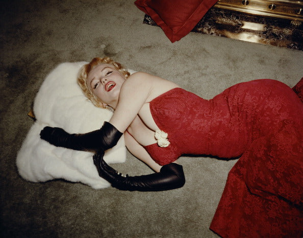 Colour photograph of Marilyn Monroe in a red brocade gown and long black gloves lying on a carpet with her head on a white fur stole, 1955