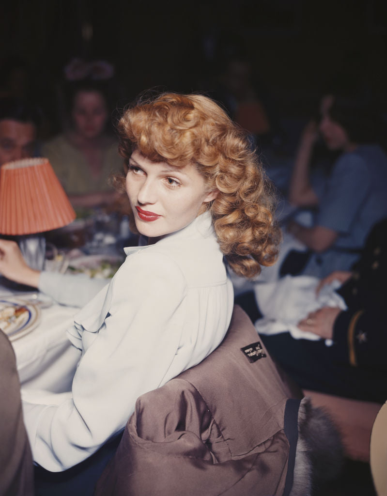 Colour photograph of Rita Hayworth with flowing red hair in a white ruffled blouse, glancing over her shoulder at a restaurant table, circa 1944