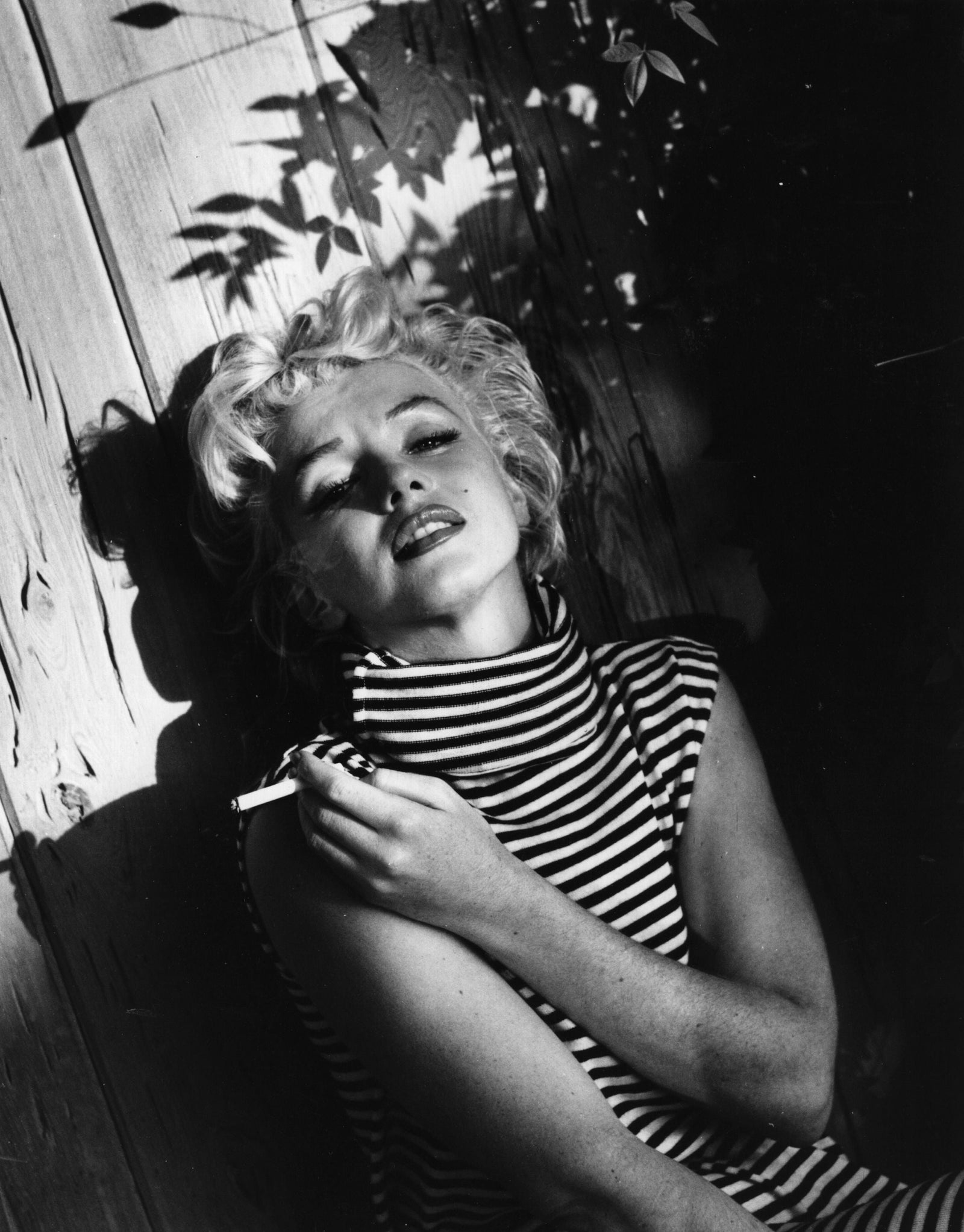 Black and white photograph of Marilyn Monroe in a striped top reclining with eyes closed and a cigarette in her garden, circa 1954