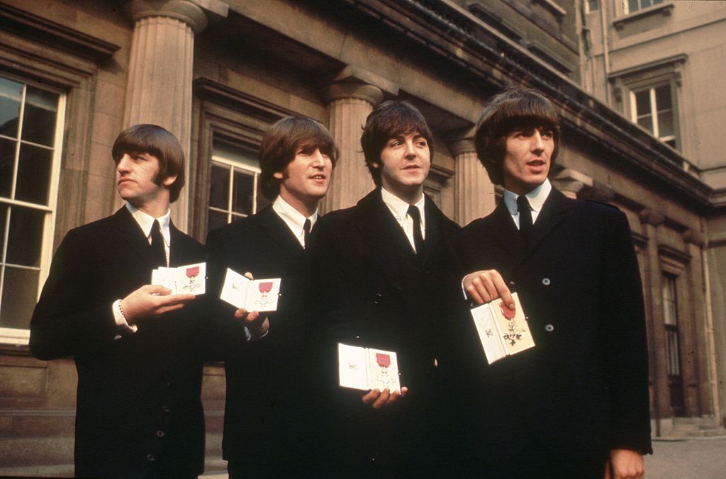 Colour photograph of The Beatles in dark suits holding their MBE medals outside Buckingham Palace, London, 1965