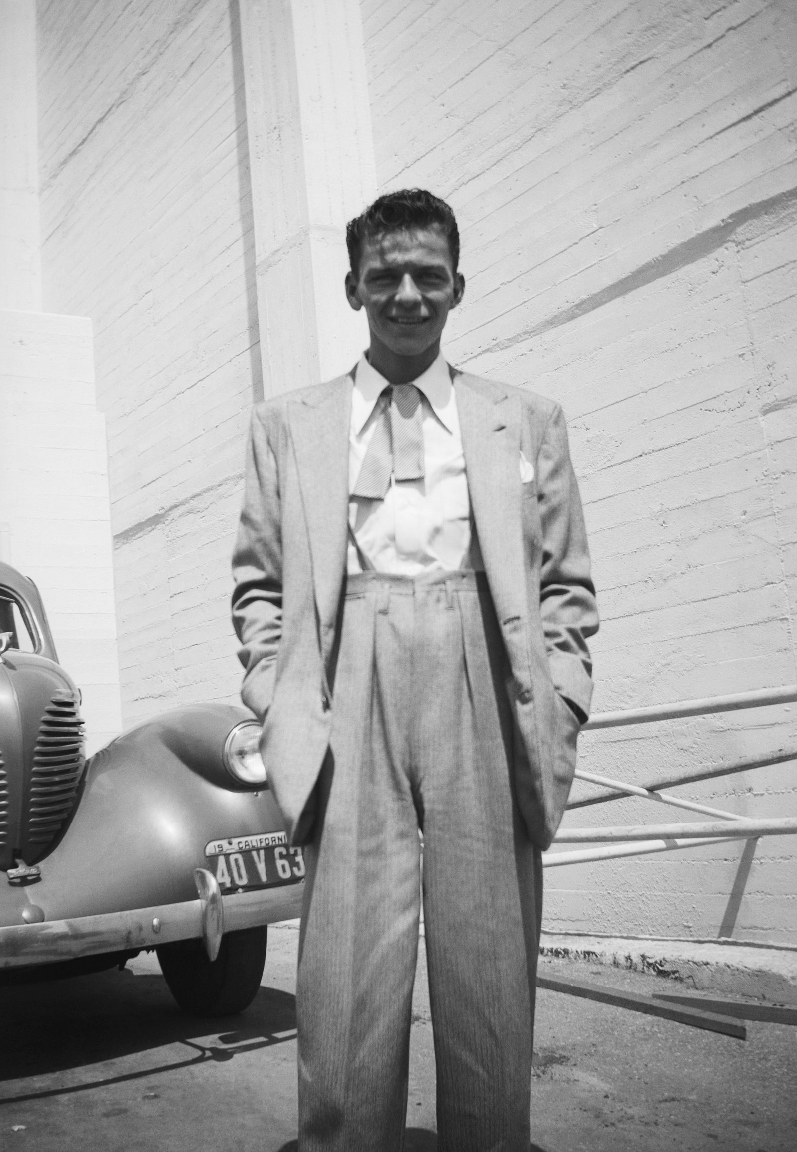 Black and white portrait of a young Frank Sinatra in a pale suit standing beside a car, Los Angeles, circa 1942