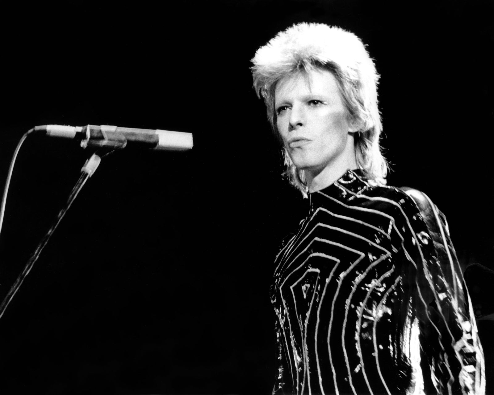 Black and white photograph of David Bowie on stage as Ziggy Stardust in a striped jumpsuit beside a microphone, Los Angeles, 1973