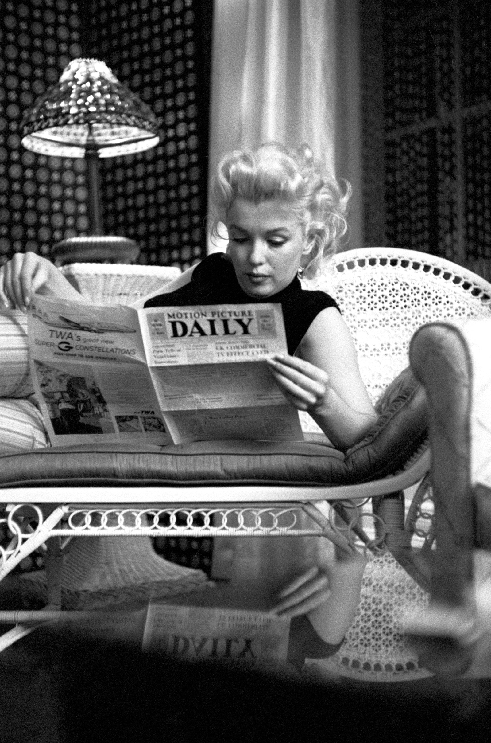 Black and white photograph of Marilyn Monroe reading Motion Picture Daily on a wicker couch at the Ambassador Hotel, New York, 1955