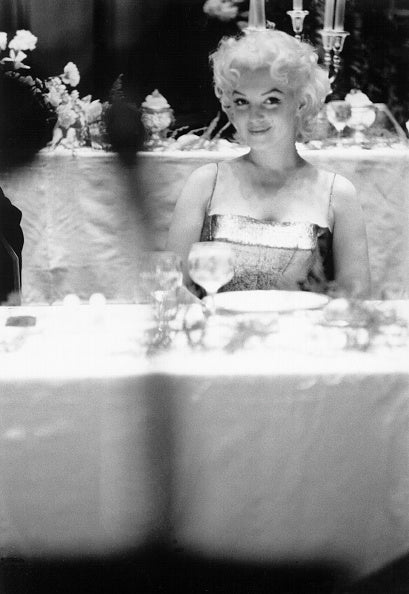 Black and white photograph of a smiling Marilyn Monroe seated alone at a white tablecloth restaurant table, New York, 1955