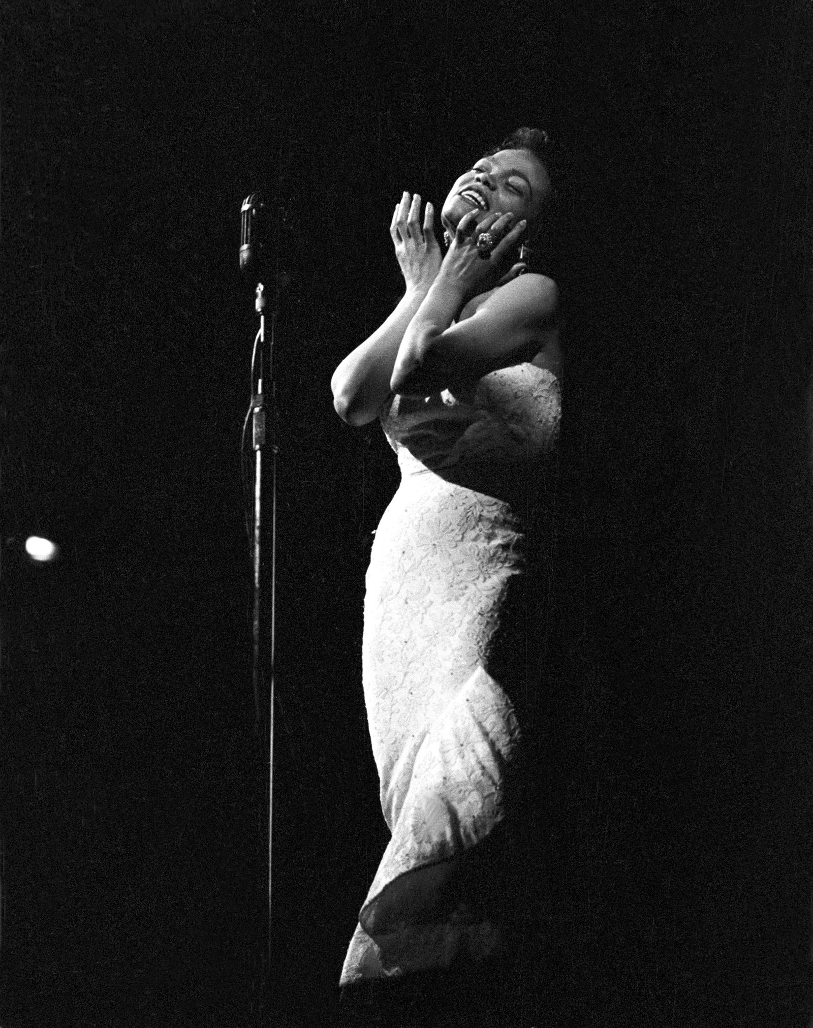 Black and white photograph of Eartha Kitt singing on a dark stage in a white sequinned gown, hands raised to her face, circa 1970