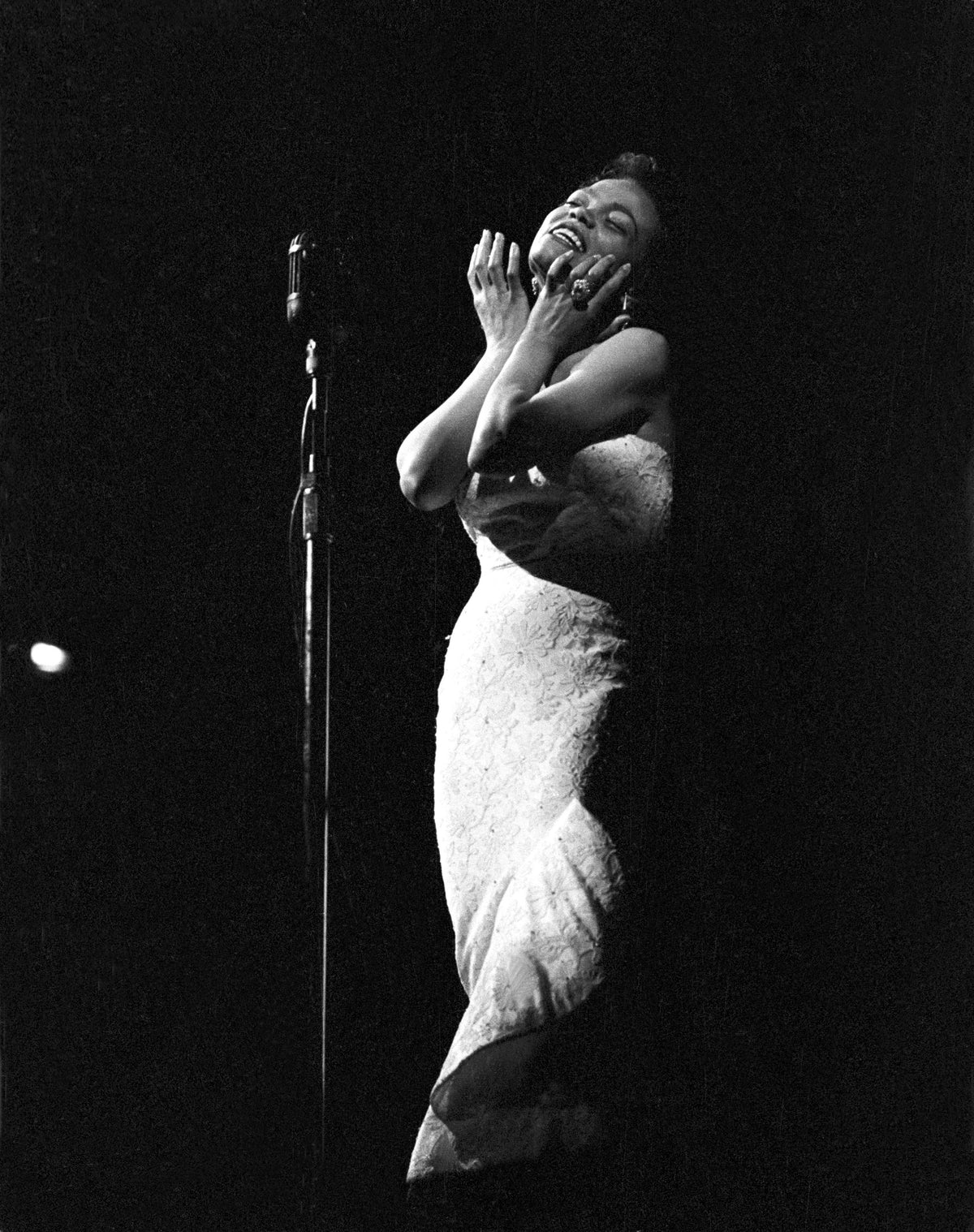 Black and white photograph of Eartha Kitt singing on a dark stage in a white sequinned gown, hands raised to her face, circa 1970