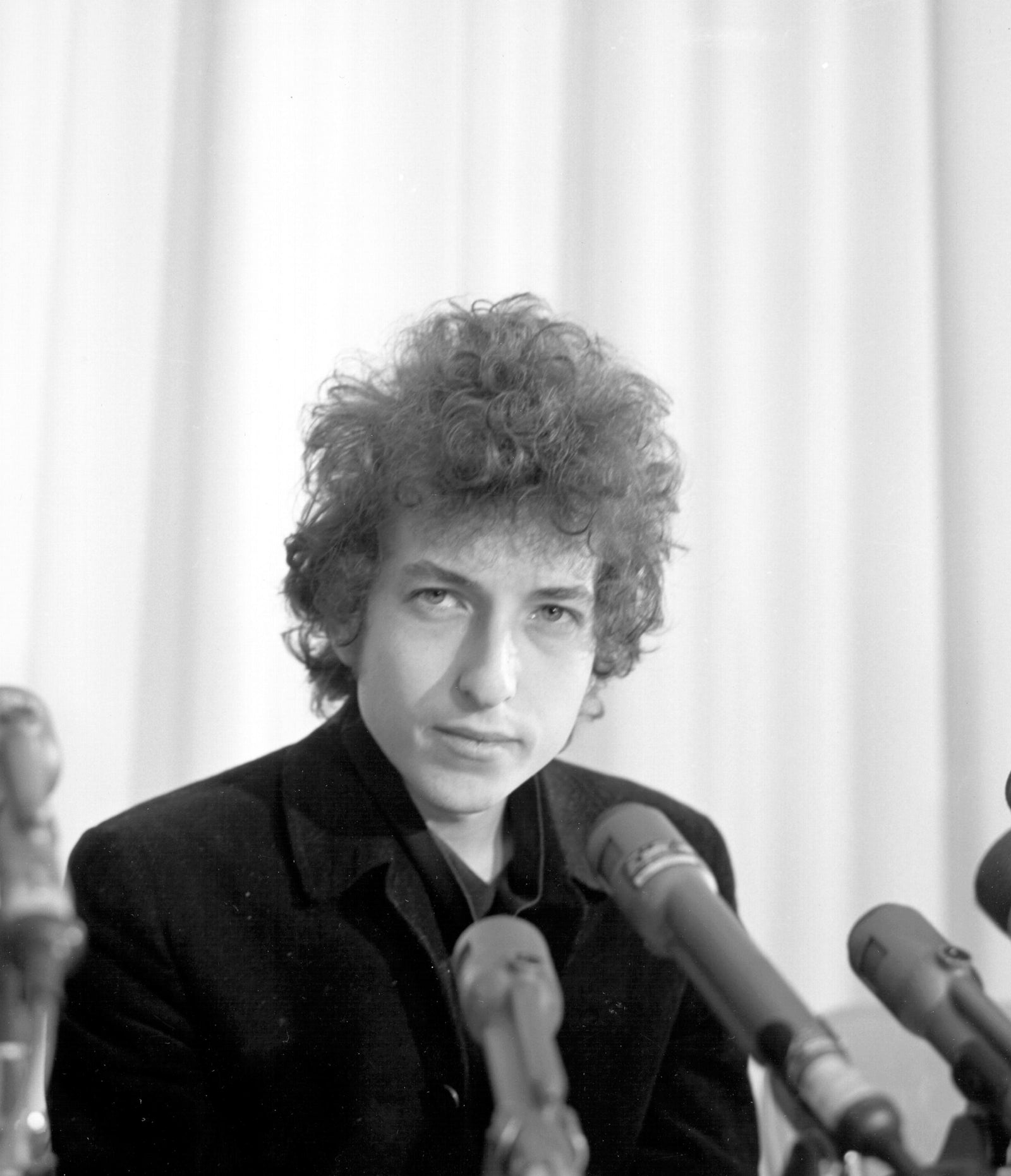Black and white photograph of Bob Dylan behind a cluster of microphones at a press conference, Los Angeles, 1965