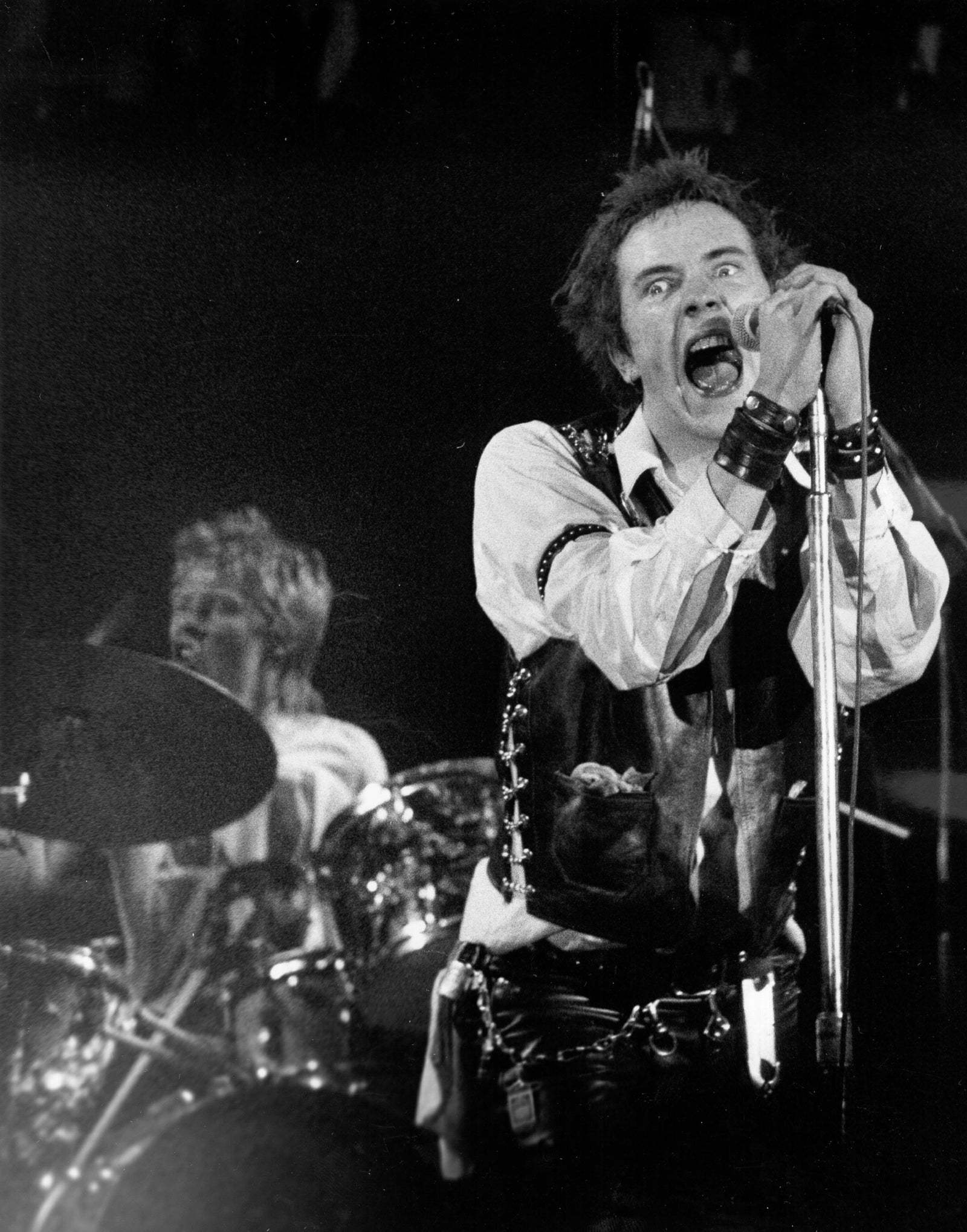 Black and white photograph of Johnny Rotten singing into a microphone with Paul Cook drumming behind at the last Sex Pistols concert, San Francisco, 1978