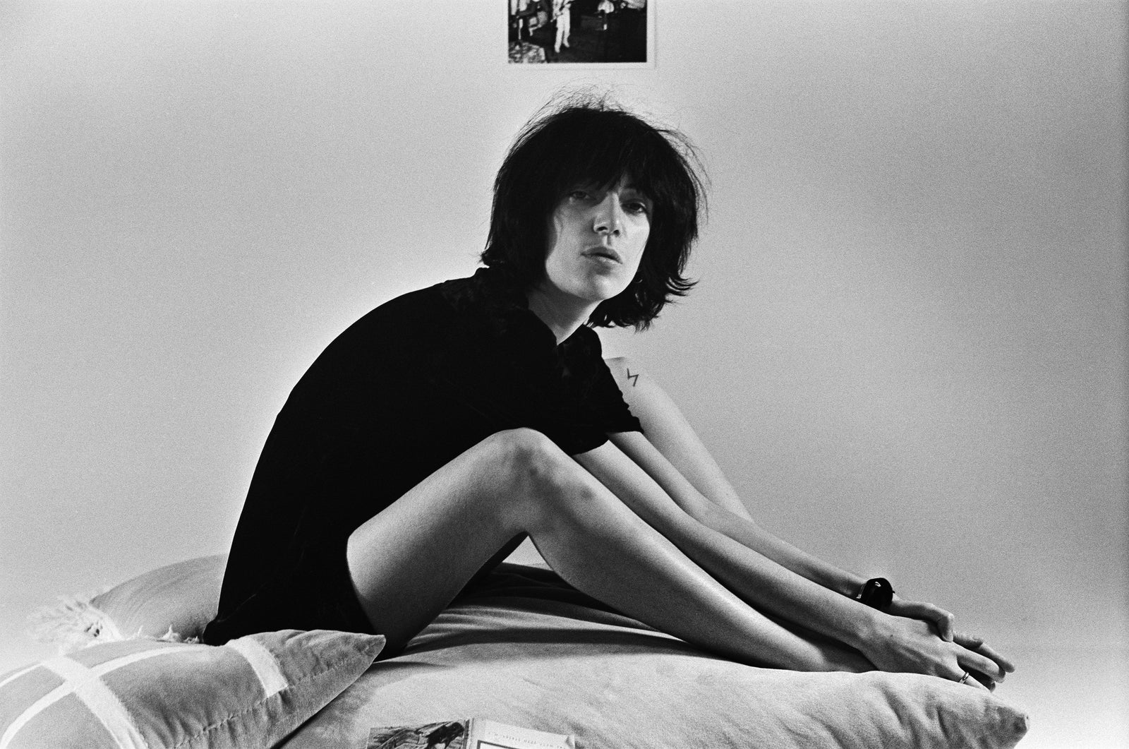 Black and white portrait of Patti Smith in a dark top sitting on a bed with her arms wrapped around her legs, 1974