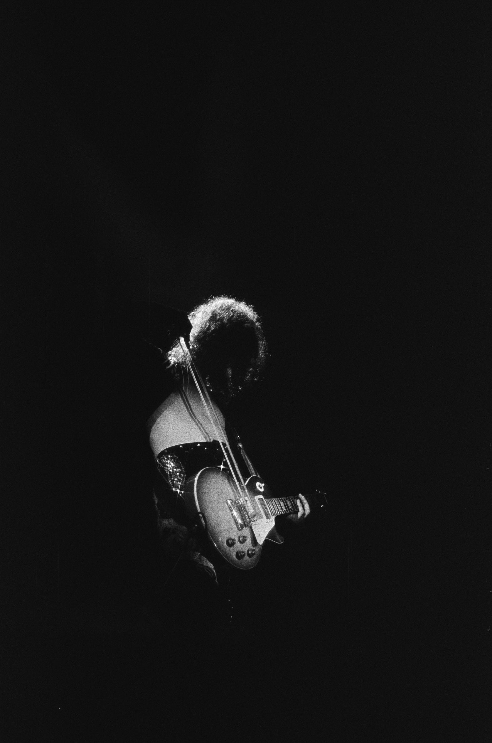 Black and white photograph of Jimmy Page playing his Les Paul guitar with a violin bow in a single spotlight at the Forum, Los Angeles, 1975