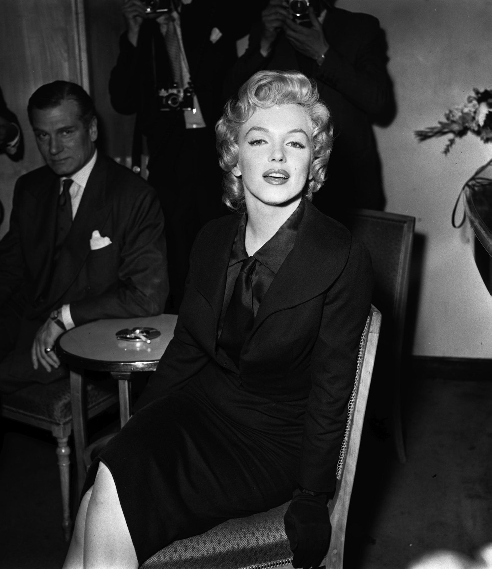 Black and white photograph of Marilyn Monroe in a dark suit seated beside Laurence Olivier at a press reception at the Savoy Hotel, London, 1956
