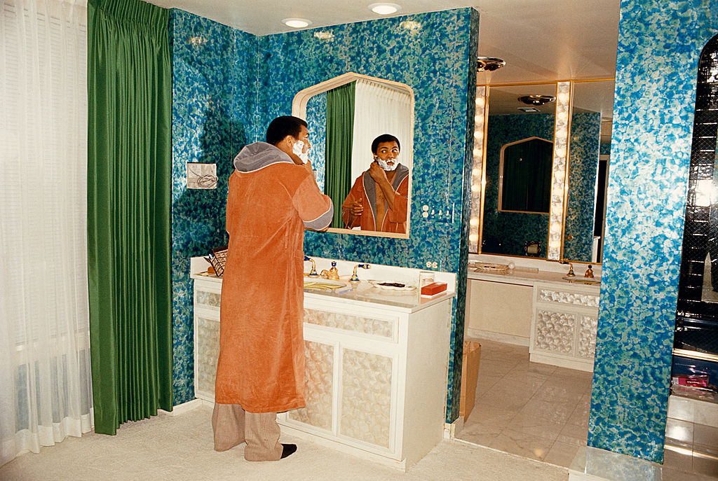 Colour photograph of Muhammad Ali in an orange robe shaving at the mirror of his blue-tiled bathroom in Chicago, 1977