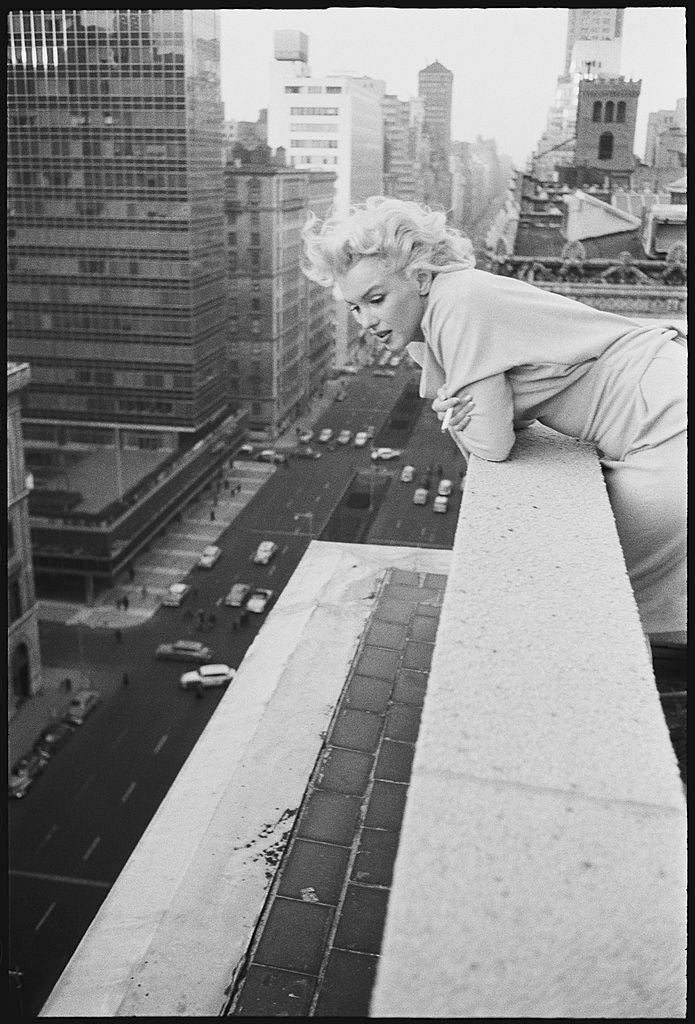 Black and white photograph of Marilyn Monroe leaning over a rooftop ledge at the Ambassador Hotel with the Manhattan skyline behind, 1955