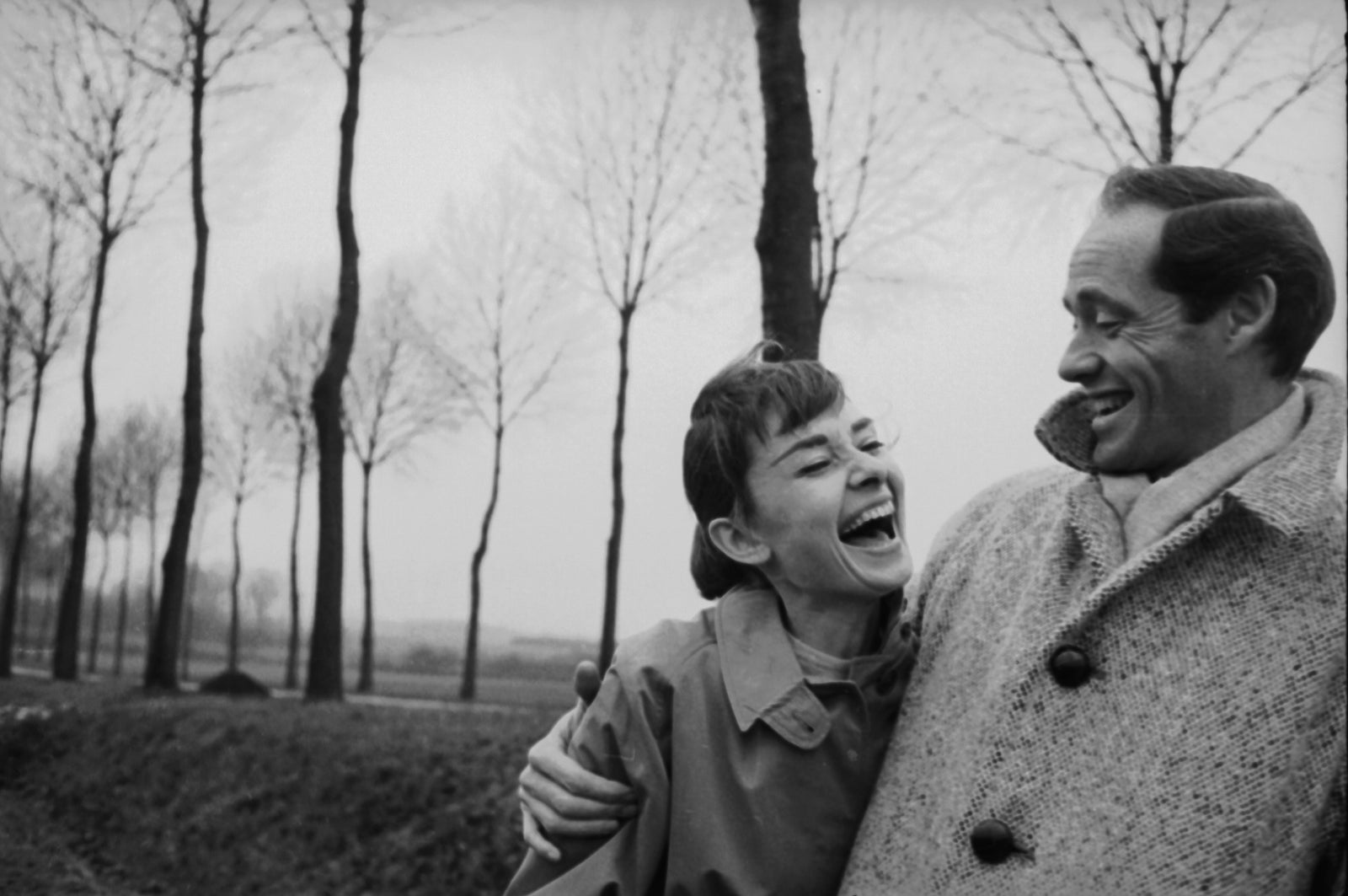 Black and white photograph of a laughing Audrey Hepburn as Mel Ferrer buttons his coat around her on a tree-lined country road near Paris, 1956