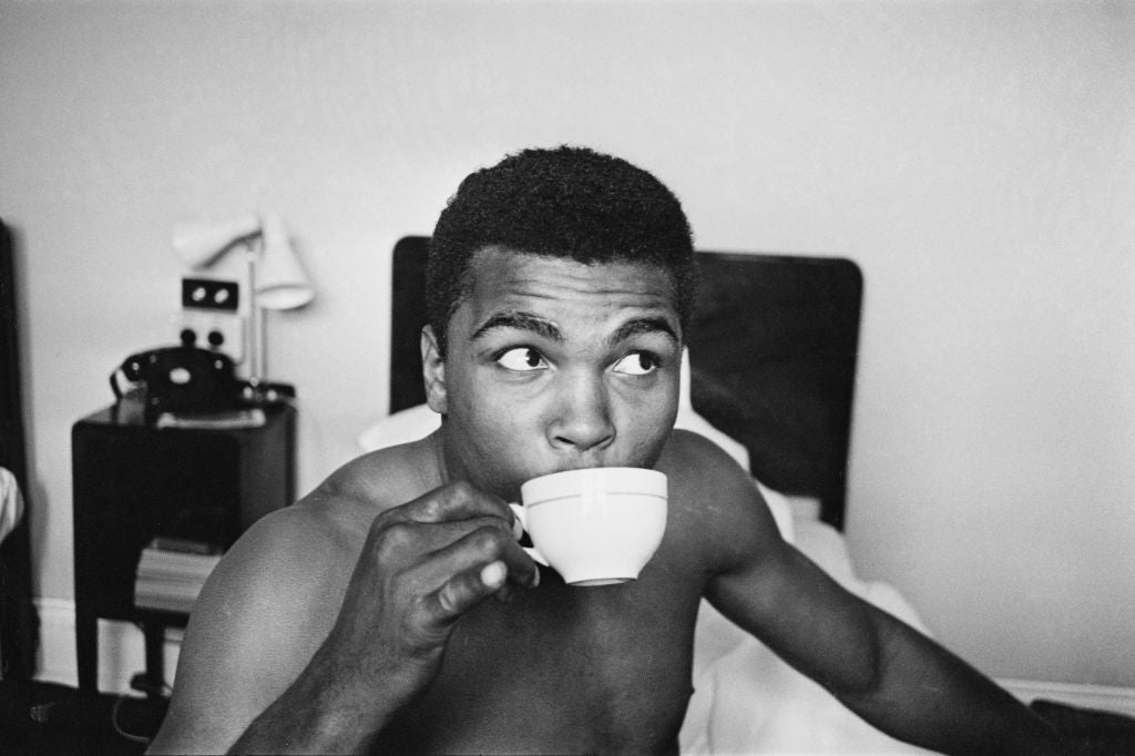 Black and white photograph of Cassius Clay sipping a cup of tea in bed in his London hotel room, 1963