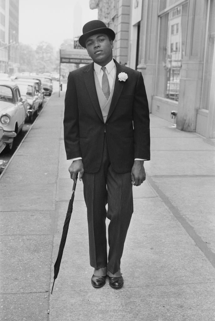 Black and white photograph of Muhammad Ali in a bowler hat, suit and carnation holding an umbrella on a New York pavement, 1963