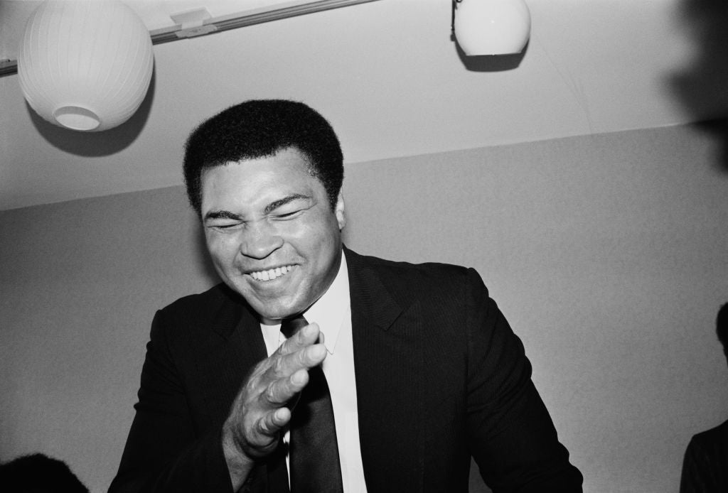 Black and white photograph of a laughing Muhammad Ali in a suit and tie gesturing towards the camera, UK, 1983