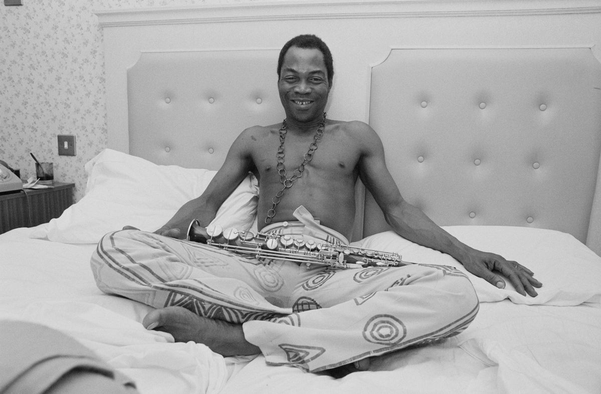 Black and white photograph of Fela Kuti sitting cross-legged and shirtless on a hotel bed, UK, 1984