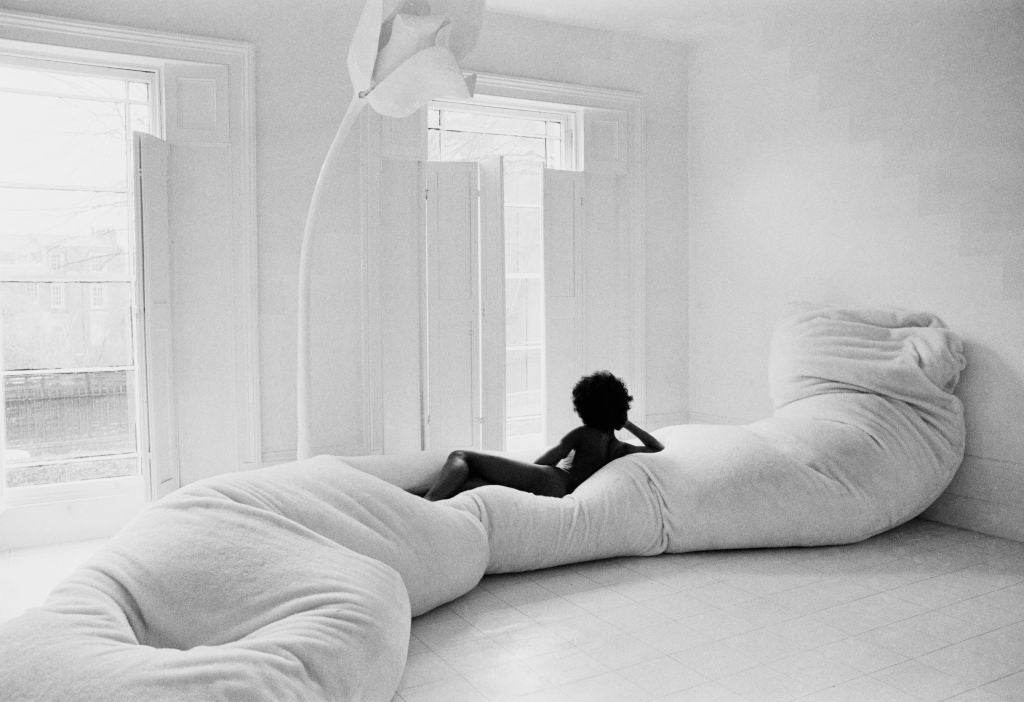 Black and white photograph of a woman lounging on a long white beanbag sofa in a minimalist all-white room, 1970