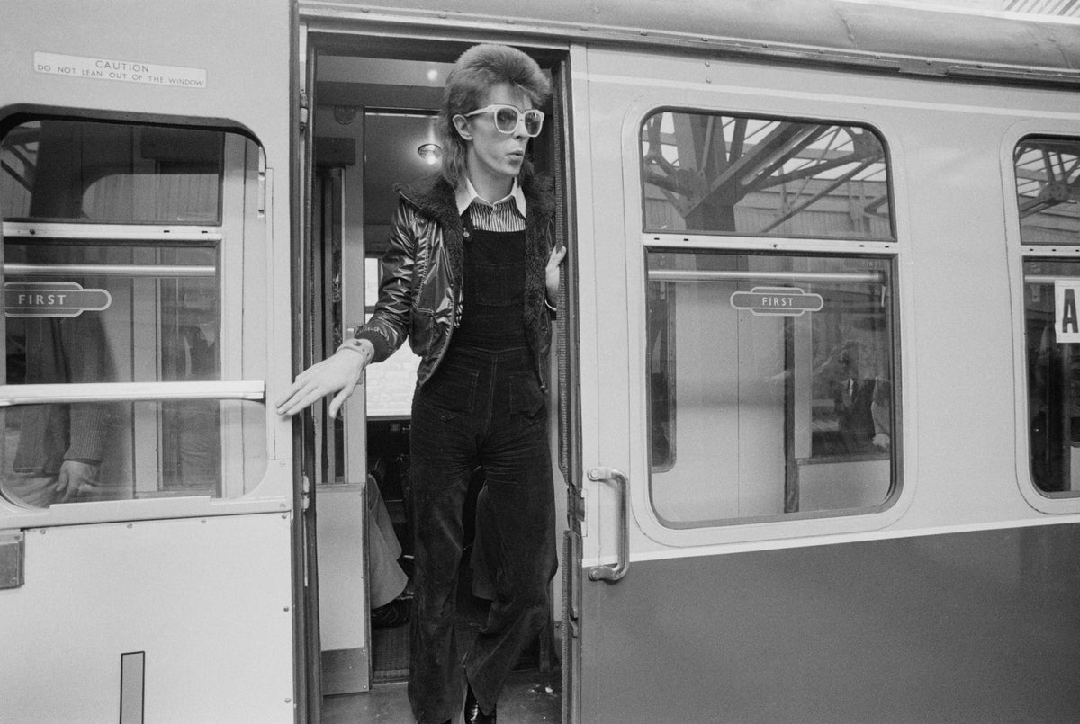 Black and white photograph of David Bowie in a jacket and dungarees stepping out of a train door at Victoria Station, London, 1973