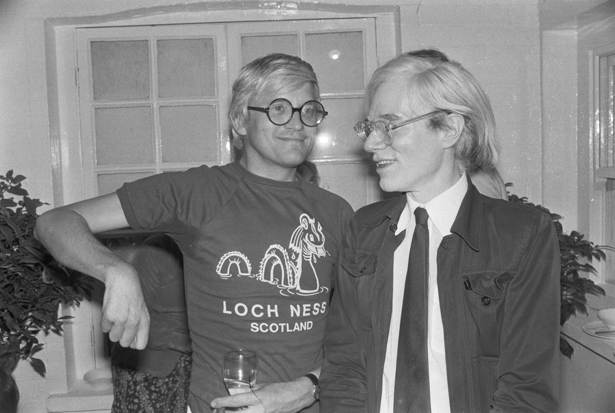 Black and white photograph of David Hockney in a Loch Ness t-shirt standing beside Andy Warhol, 1976