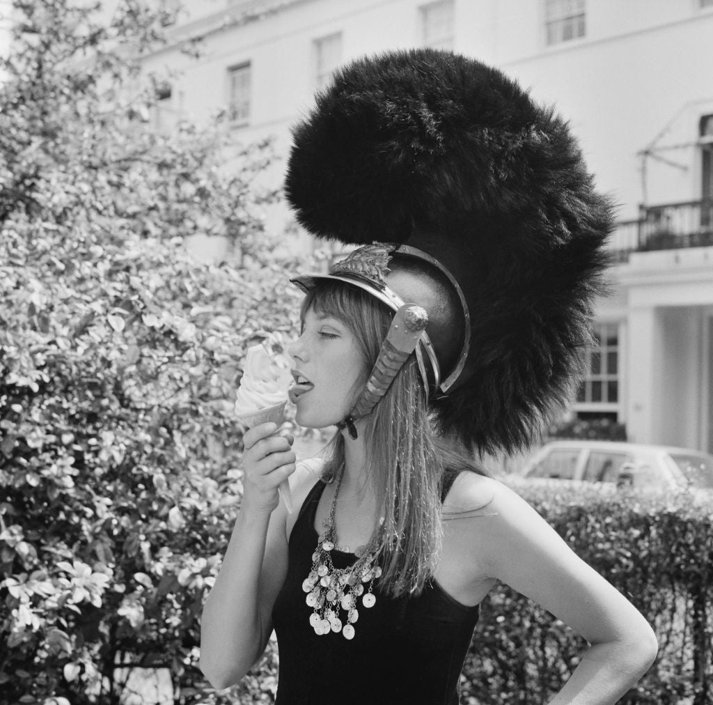 Black and white photograph of Jane Birkin eating an ice cream while wearing a plumed military helmet on a leafy London street, 1970