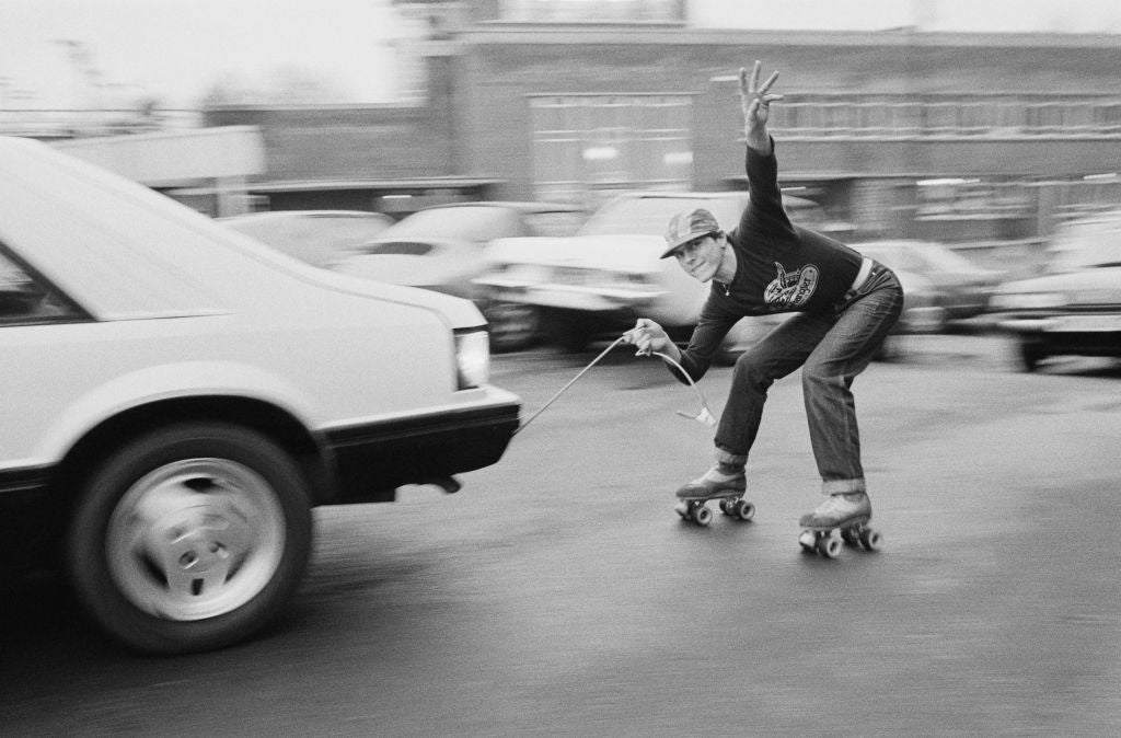 Black and white photograph of roller skater Leo Bull crouched low on quad skates, towed by a cable behind a car, 1980