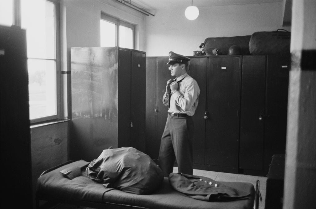 Black and white photograph of Elvis Presley in US Army uniform standing by lockers in a barracks room, 1958