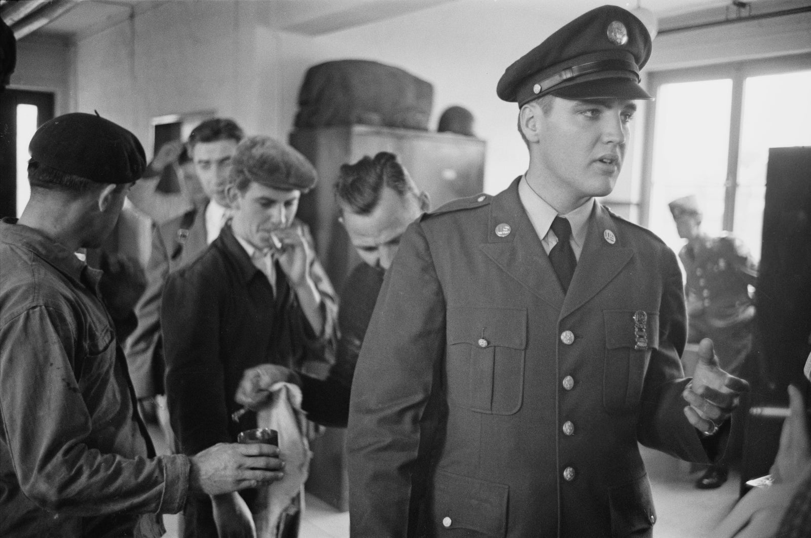 Black and white photograph of Elvis Presley in US Army uniform and cap surrounded by fellow soldiers, 1958