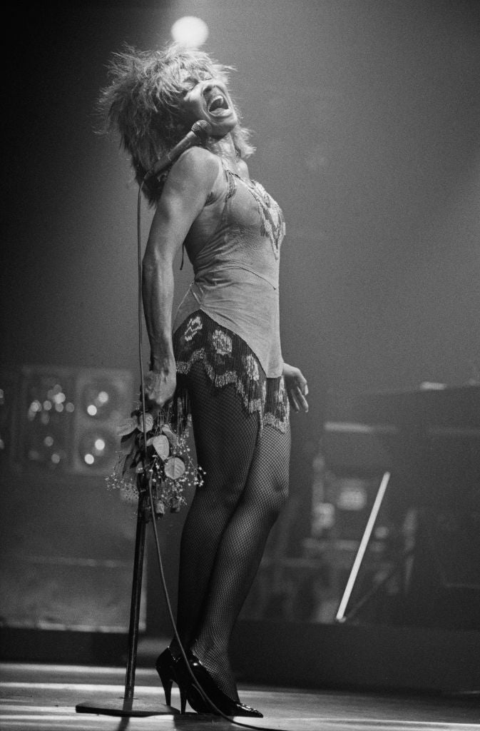 Black and white photograph of Tina Turner performing on stage at the Brighton Centre, head thrown back beside a microphone stand, 1985