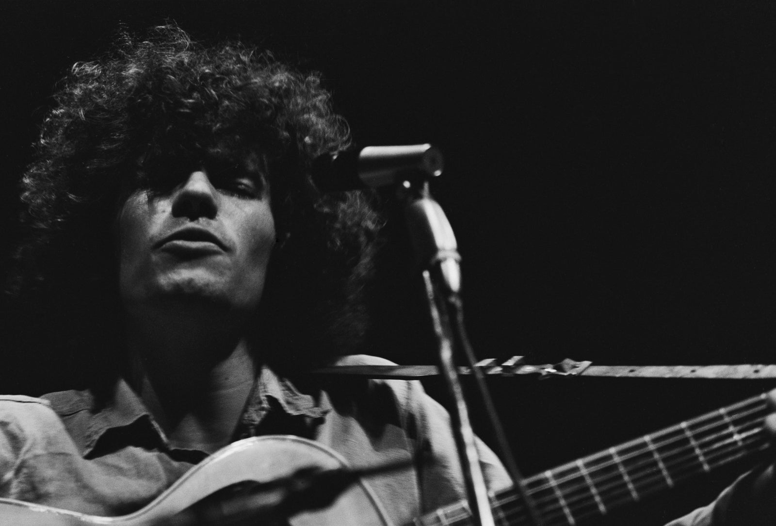 Black and white close-up of Tim Buckley singing at a microphone with his acoustic guitar, circa 1967