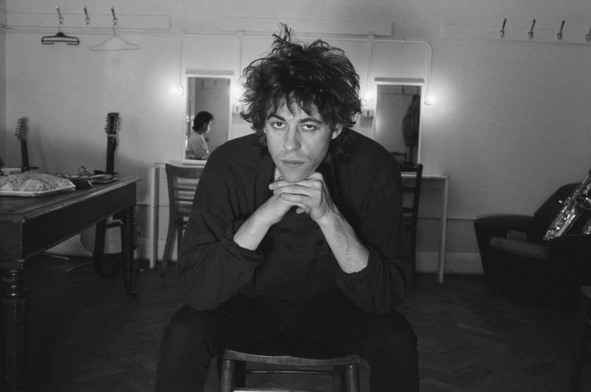 Black and white photograph of Bob Geldof seated in a dressing room, chin resting on his clasped hands, 1985