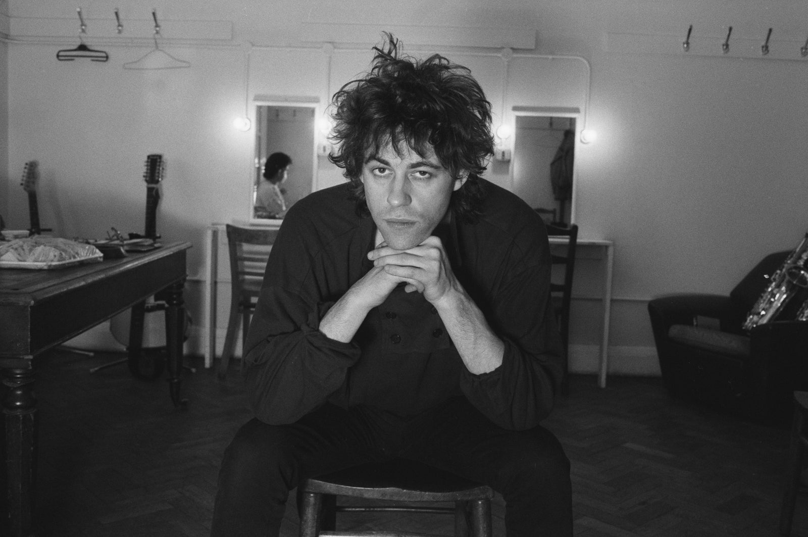 Black and white photograph of Bob Geldof seated in a dressing room, chin resting on his clasped hands, 1985